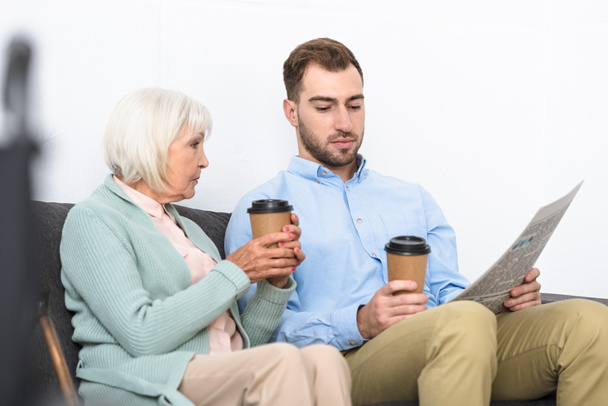 man en senior moeder houden koffie en lezen krant thuis - Foto, afbeelding