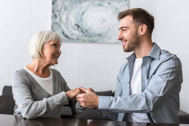 smiling man holding hands with senior mother a home - Foto, Bild