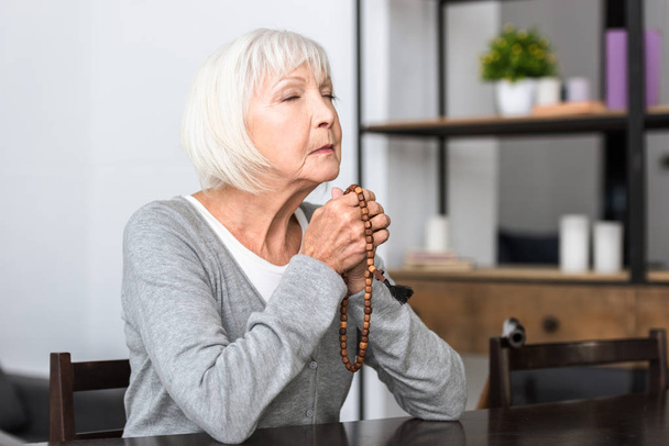 Seniorin hält hölzernen Rosenkranz und betet mit geschlossenen Augen - Foto, Bild