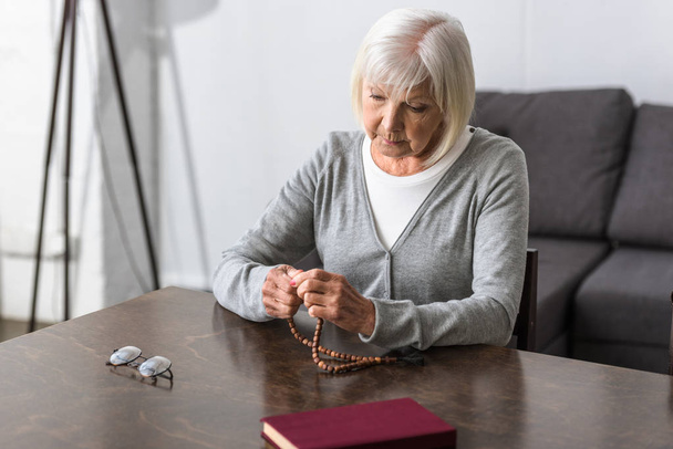 Nachdenkliche Seniorin sitzt am Tisch und hält Rosenkranz in der Hand - Foto, Bild