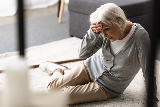Senior vrouw met migraine zittend op tapijt en aanraken van voorhoofd met de hand - Foto, afbeelding