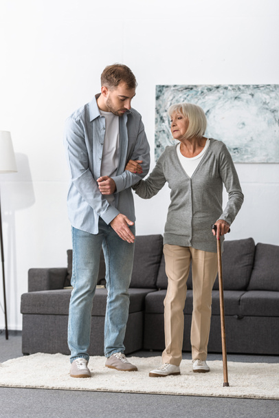 full length view of man helping senior mother with cane at home - Foto, afbeelding