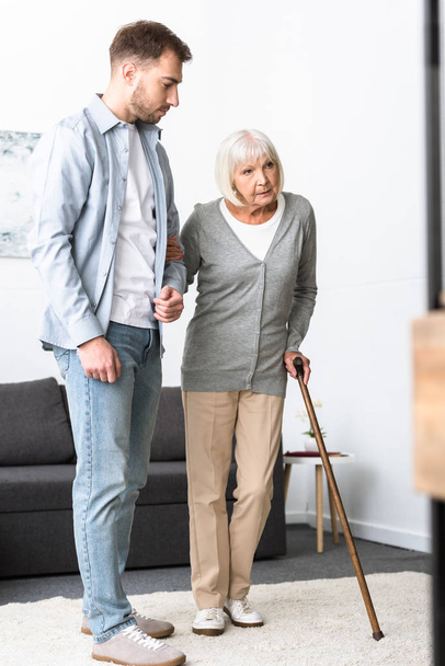 full length view of man helping senior mother with cane at home - Foto, afbeelding