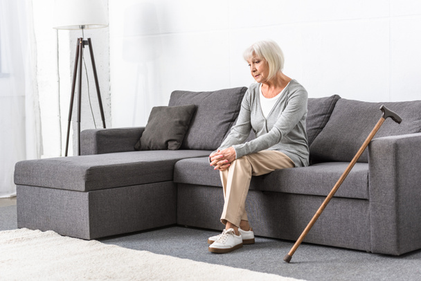 pensive senior woman with wooden cane sitting on sofa in living room - Photo, Image