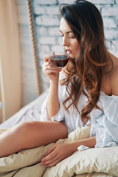 Young beautiful woman drinking hot coffee sitting in bed and enjoying her good morning. Relax and carefrre concept - Photo, Image