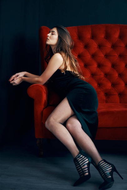 Portrait of sexy sensual woman in black dress posing on red sofa. Fashion concept                                - Фото, зображення
