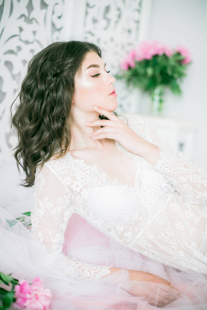 Beautiful young girl in a white lace dress with peony flowers in a bright studio - Photo, image