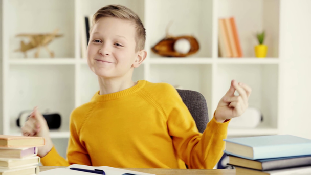 criança feliz estalando os dedos enquanto sentado na cadeira perto de livros, sorrindo e tomando caneta
  - Filmagem, Vídeo