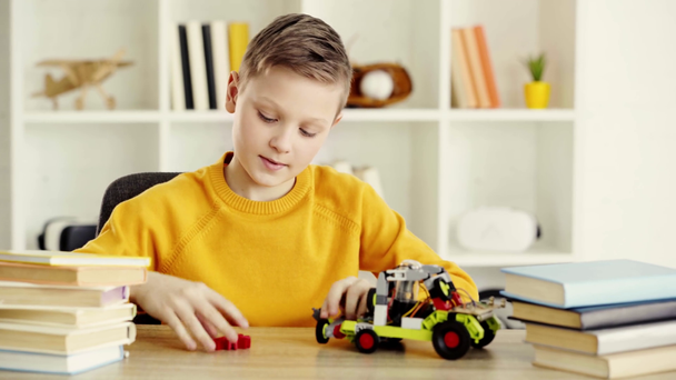 szczęśliwy dzieciak bawiąc się samochód elektryczny wykonany z bloków konstrukcyjnych w pobliżu książek na stole w domu  - Materiał filmowy, wideo
