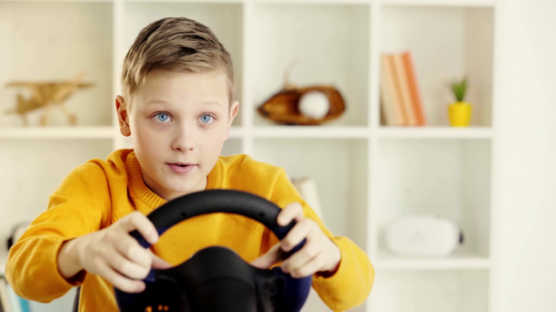 επιλεκτική εστίαση του συναισθηματικού παιδιού κρατώντας το τιμόνι παιχνίδι και παίζοντας βίντεο παιχνίδι στο σπίτι  - Πλάνα, βίντεο