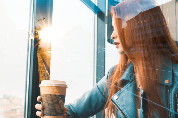 beautiful romantic girl drinks coffee in a cafe. red-haired woman sitting near the window. against the backdrop of the city grows in the sunset rays - Foto, Bild