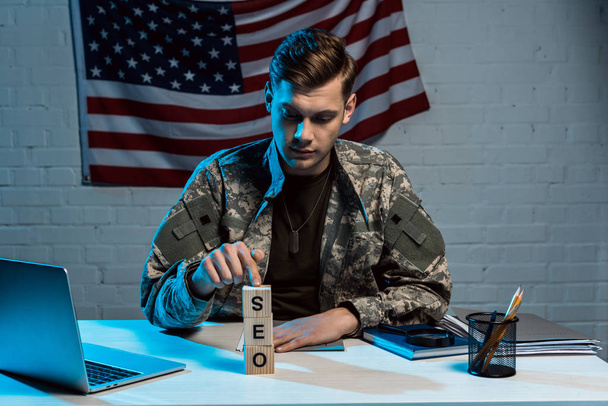 handsome military man sitting near laptop and pointing with finger at cubes with seo lettering  - Foto, imagen