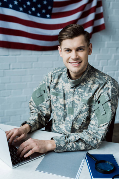 веселый солдат смотрит в камеру во время использования ноутбука в офисе
  - Фото, изображение