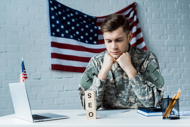 rozčilený voják, který se dívá na dřevěné kostky s písmem SEO v kanceláři  - Fotografie, Obrázek