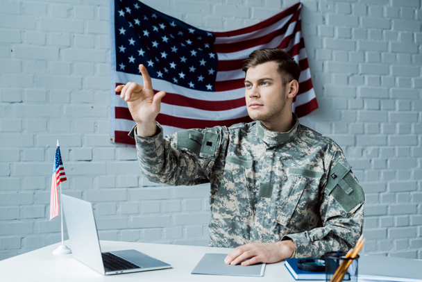 красивый мужчина в военной форме указывает пальцем и сидит рядом с ноутбуком
  - Фото, изображение