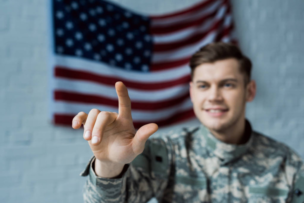 Amerikan bayrağı yakınında parmak ile işaret askeri üniformalı mutlu adam seçici odak - Fotoğraf, Görsel