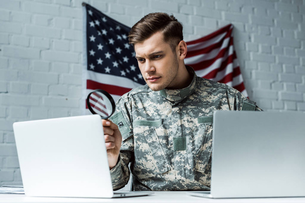 όμορφος στρατιώτης κρατώντας μεγεθυντικό φακό κοντά σε φορητούς υπολογιστές  - Φωτογραφία, εικόνα
