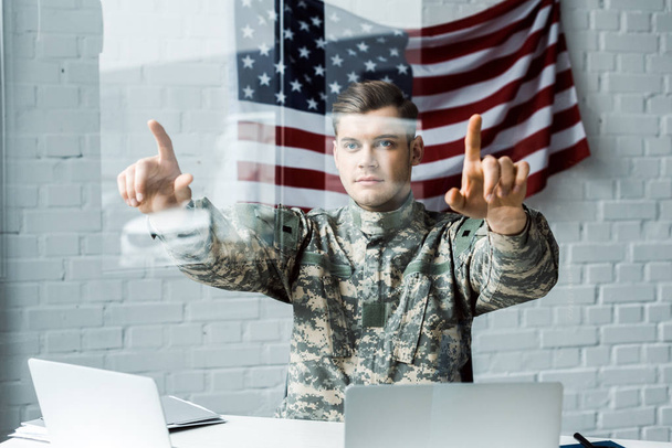 επιλεκτική εστίαση του όμορφου στρατιώτη σε ομοιόμορφη δείχνοντας με τα δάχτυλα κοντά σε φορητούς υπολογιστές στο γραφείο  - Φωτογραφία, εικόνα