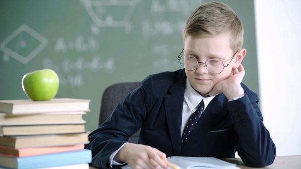 学校で親指を見せながら、本やリンゴの近くにノートに書き込むメガネの悲しい男子生徒  - 映像、動画