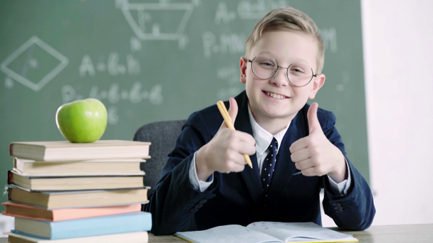 happy schoolboy in glasses writing in notebook near books and apple, while showing thumbs up in school  - Footage, Video
