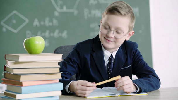 school jongen in glazen schrijven in notebook, knikkend en glimlachend in de buurt van boeken en groene appel op school  - Video