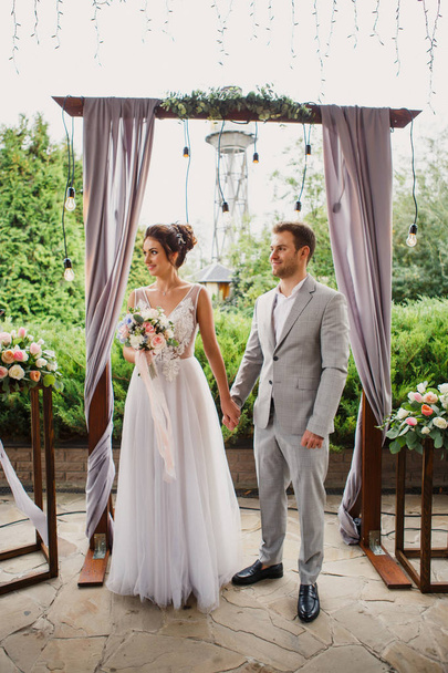 Beautiful wedding couple. The bride and groom are standing at the wedding ceremony. The bride in a beautiful dress he groom is dressed stylishly - Zdjęcie, obraz