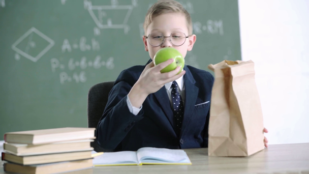 šťastný školák v brýlích, který si bere papírový pytel a dívá se na zelený jablíčko, zatímco ve škole ukazoval palec  - Záběry, video