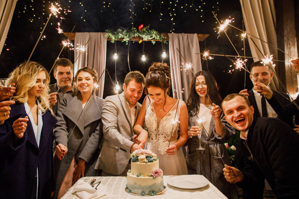 若い美しい結婚式のカップルは友人と結婚式のケーキをカットし、優しい照明で楽しみます - 写真・画像