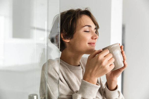 Blij brunette vrouw drinken koffie met gesloten ogen en geniet - Foto, afbeelding