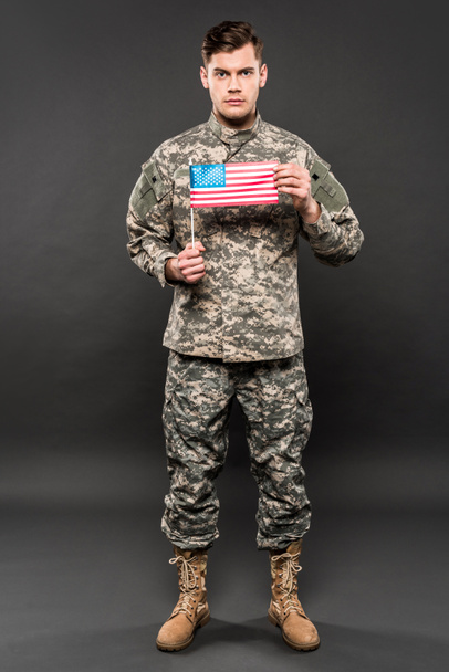serious man in military uniform holding american flag on grey  - Φωτογραφία, εικόνα
