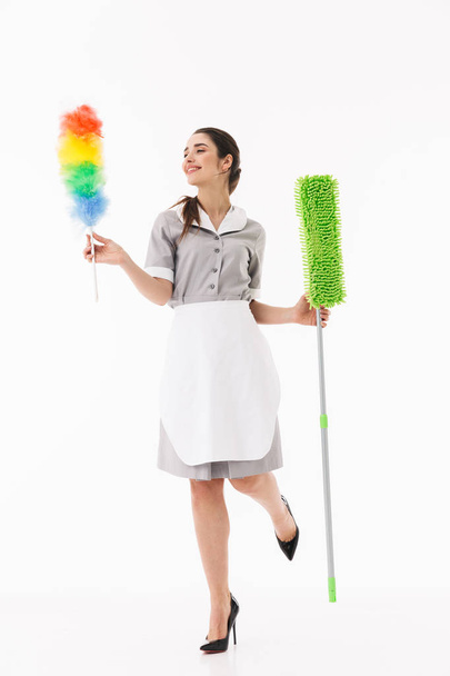 Full length photo of caucasian maid 20s dressed in uniform holding colorful duster and mop while doing housework isolated over white background - Photo, image