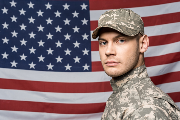 handsome man in military uniform and cap looking at camera near flag of america - Фото, зображення