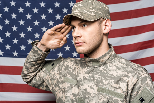 handsome soldier in military uniform and cap giving salute near flag of america - Photo, Image