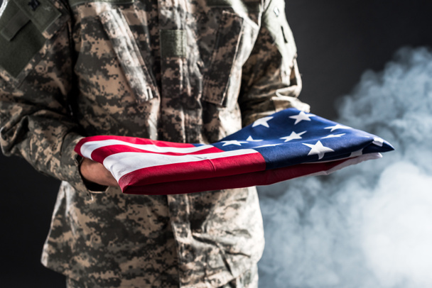bijgesneden beeld van de mens in militaire uniforme Holding vlag van Amerika op zwart met rook  - Foto, afbeelding