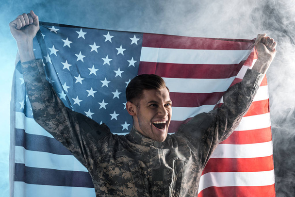 happy soldier gesturing while holding american flag on black with smoke  - Photo, Image