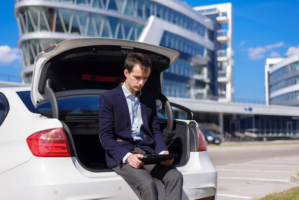 successful young businessman freelancer working outside in the city. man sit on car boot with tablet  - Photo, Image