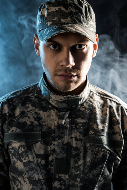 confident soldier in military cap and uniform looking at camera on black with smoke - Fotografie, Obrázek