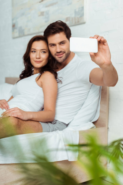 selective focus of handsome man taking selfie with attractive girlfriend in bedroom  - Fotoğraf, Görsel