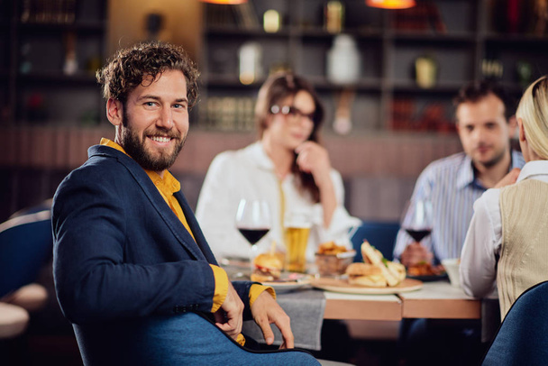 Young smiling man dressed smart casual sitting at restaurant and looking over shoulder. In background his friends having dinner. - Fotoğraf, Görsel