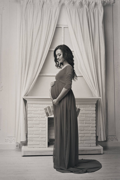 красивая беременная девушка в длинном платье, прикасаясь к животу на фоне домашнего интерьера: камин и шторы
 - Фото, изображение