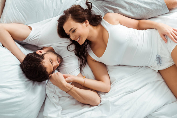 top view of cheerful woman looking at man while holding hands and lying on bed  - Фото, изображение