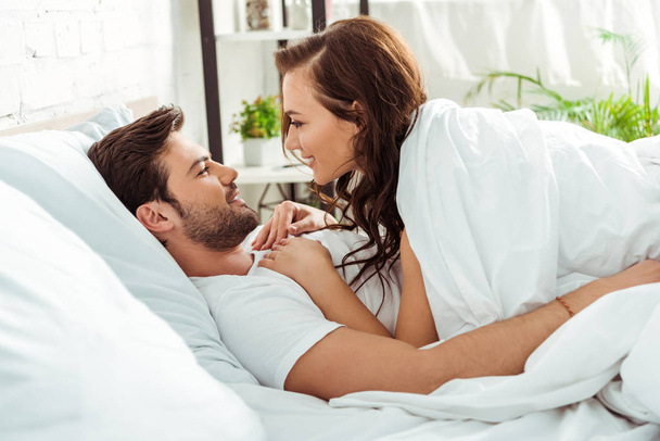 happy woman looking at smiling boyfriend while lying in bedroom  - Photo, Image