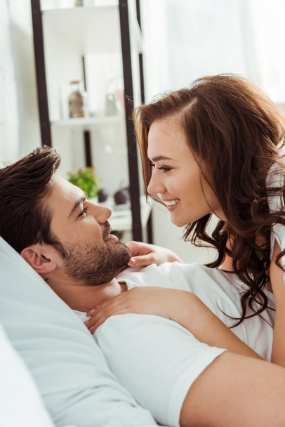 happy woman looking at handsome boyfriend while lying in bedroom  - Фото, изображение