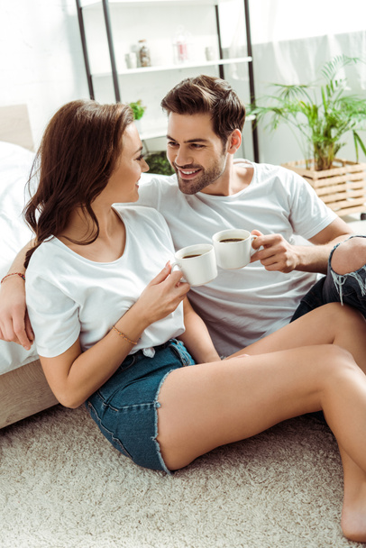 handsome man and cheerful woman holding cups of coffee in bedroom  - Fotó, kép