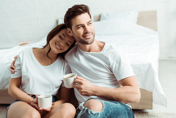 happy man sitting with cheerful girl and holding cup in bedroom  - Foto, afbeelding