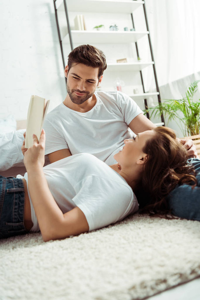 low angle view of handsome man looking at book in hands of girlfriend lying on carpet in bedroom  - Fotó, kép