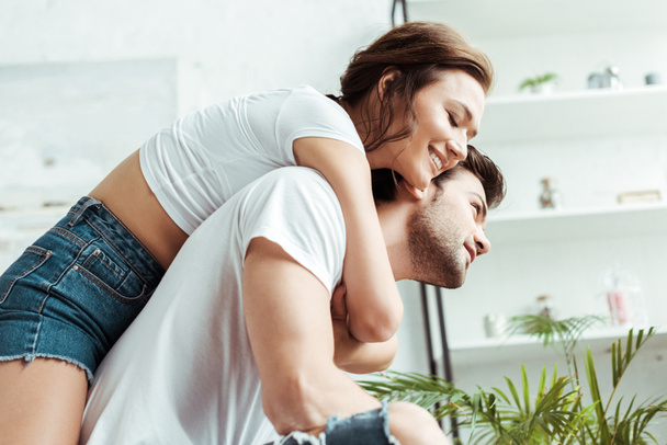 happy girl hugging handsome boyfriend in white t-shirt and smiling at home  - Фото, изображение