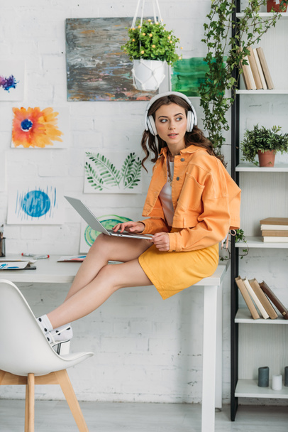 mujer joven de moda en los auriculares usando el ordenador portátil mientras está sentado en el escritorio en casa
 - Foto, Imagen