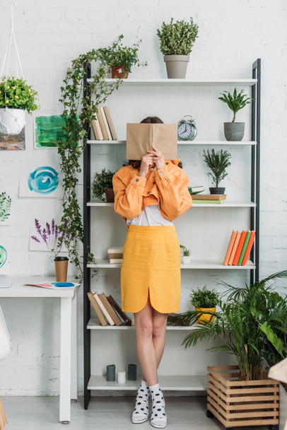 fashionable young woman standing bu rack and hiding face behind book - Foto, afbeelding