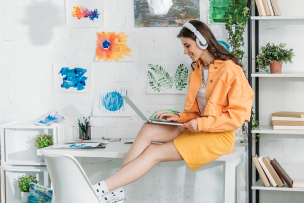 smiling trendy woman in headphones using laptop while sitting on desk - 写真・画像